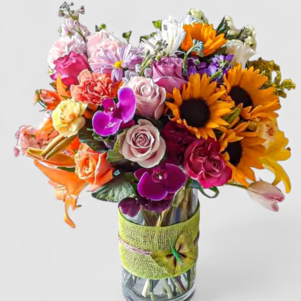 Bright mixed bouquet of sunflowers, roses, lilies, and orchids in a glass vase with green wrap.
