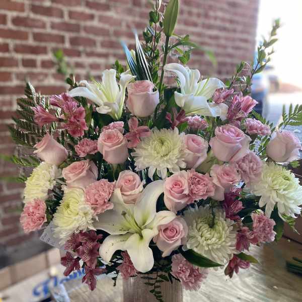 Arrangement of pink roses, carnations, and white lilies in a textured vase on a wooden table.