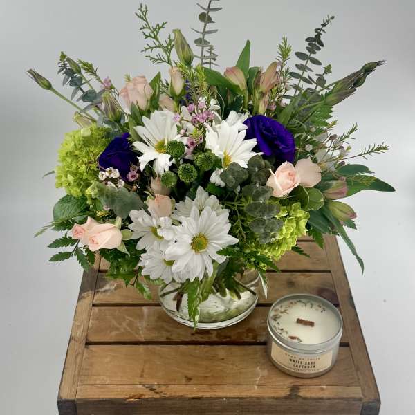 Mixed bouquet of white daisies, pink roses, and purple blooms in a glass bowl with a candle on a wooden crate