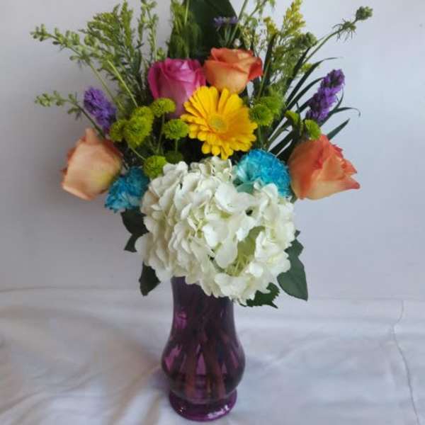 Tall mixed arrangement with roses, gerbera, hydrangea and mums in a purple glass vase