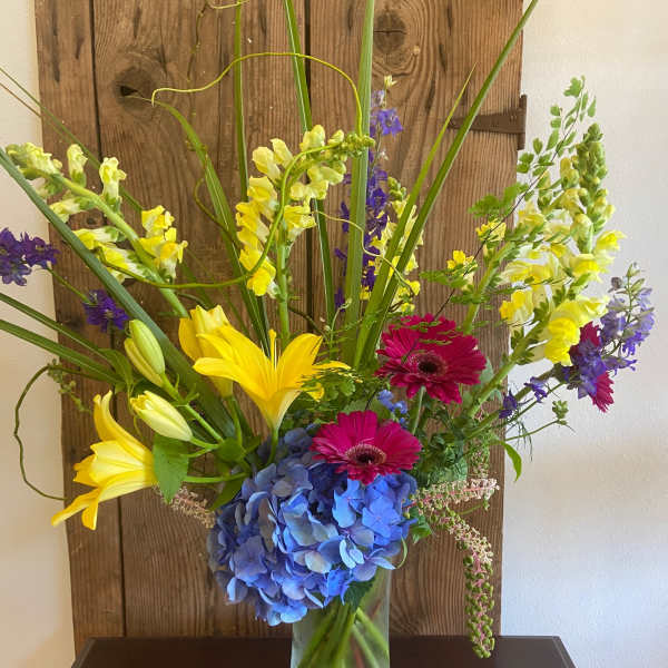 Tall mixed arrangement with yellow lilies, snapdragons, pink gerberas and blue hydrangeas in a clear glass vase