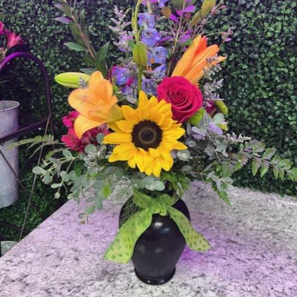 Tall mixed bouquet with sunflower, lilies, rose, and blue delphinium in a black vase with green bow.