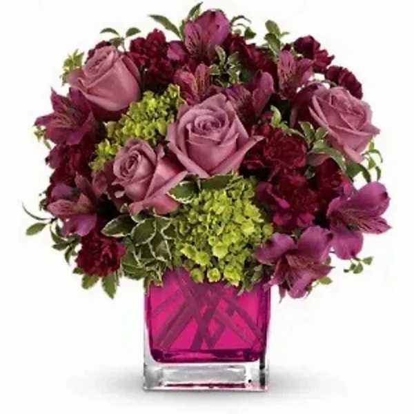 Compact arrangement of mauve roses, dark carnations, and green hydrangea in a bright pink glass cube vase