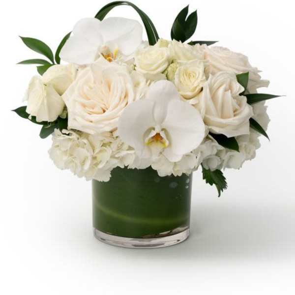 Compact arrangement of white roses, hydrangeas, and orchids in a low glass vase with green lining