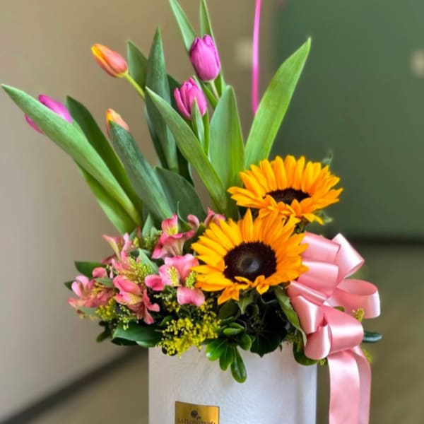 Bright arrangement with a sunflower, pink tulips, and pink blooms in a white hatbox with pink ribbon bow