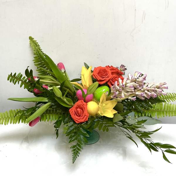 Low arrangement with orange roses, yellow lilies, pink tulips and pastel hyacinth with colorful eggs in a green vase