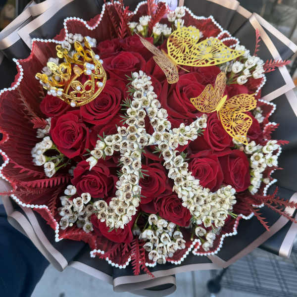 Hand-tied bouquet of red roses with white flowers shaped into a letter and gold crown and butterfly accents