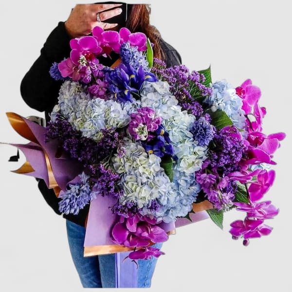 Large hand-tied bouquet of purple orchids, blue hydrangeas, and mixed purple flowers in pastel wrap.