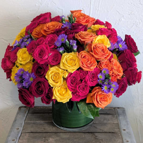 Bright arrangement of pink, yellow, red, and orange roses with purple asters in a leaf-lined glass vase.