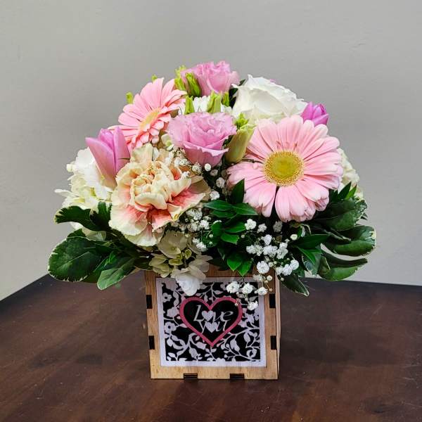 Short arrangement of pink and white flowers in a wooden cube with a heart that reads love you.