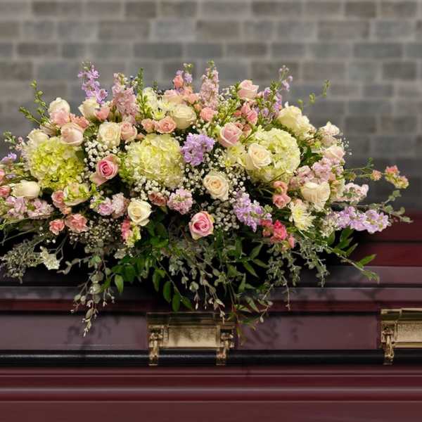 Pastel pink and cream casket spray with roses and hydrangeas on a dark wood casket