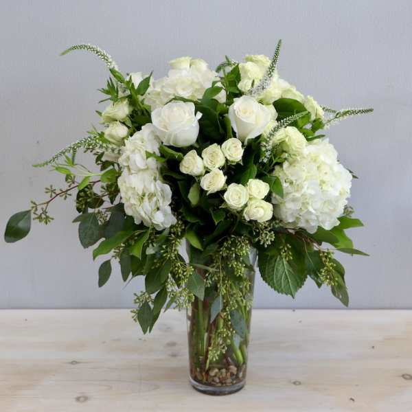 Tall arrangement of white roses and hydrangeas with trailing greenery in a clear glass vase.