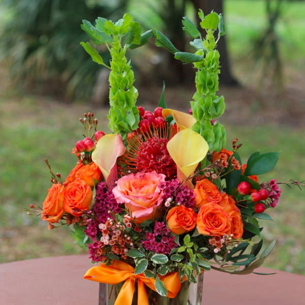 Bright orange and pink rose arrangement with yellow calla lilies and tall green bells of Ireland in a glass vase