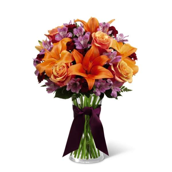 Vase arrangement of orange lilies, peach roses, and purple flowers in a clear glass vase with dark ribbon