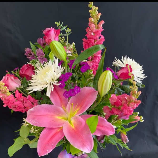 Tall mixed arrangement of pink lilies, roses, snapdragons and white mums in a clear vase with a ribbon bow