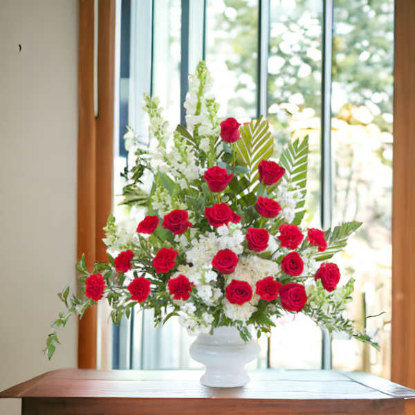 Tall arrangement of red roses and carnations with white flowers in a white urn on a table