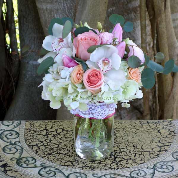 Mason jar arrangement with pink and peach roses, white orchids, hydrangeas, and pink lilies.