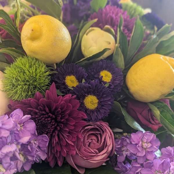 Arrangement of purple flowers with lemons and green pom blooms in a glass vase