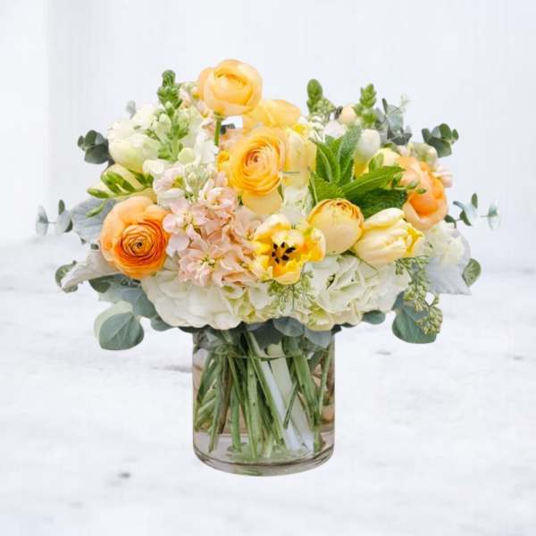 Yellow and peach ranunculus, tulips, and white hydrangeas arranged in a clear glass vase
