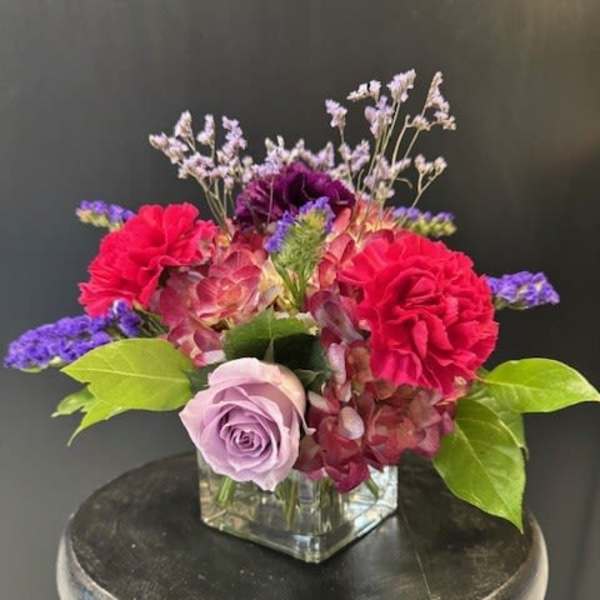 Low arrangement of pink and purple flowers with a lavender rose in a square glass vase.