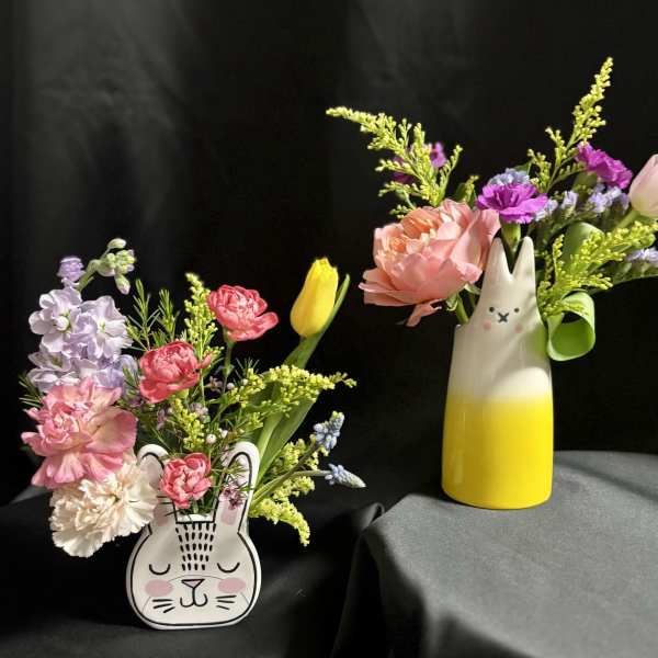 Two spring flower arrangements in bunny-shaped ceramic vases with pink, yellow, and purple blooms on a dark background
