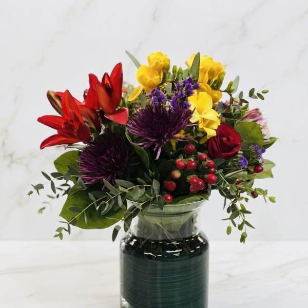 Bright mixed bouquet with red lilies, yellow blooms, purple mums and red roses in a clear glass vase