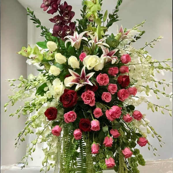 Tall standing floral spray with red and pink roses, lilies, orchids and trailing greenery.