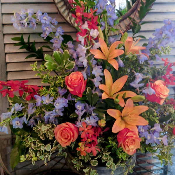 Tall arrangement of peach lilies, coral roses, blue delphinium and red orchids in a metal container