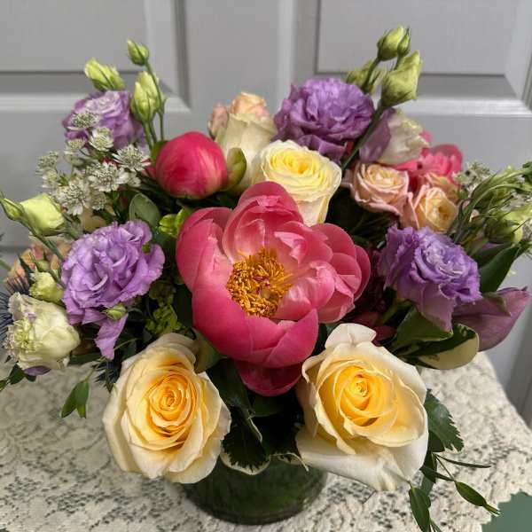 Low mixed bouquet of pink peonies, cream roses, and purple lisianthus in a clear glass vase