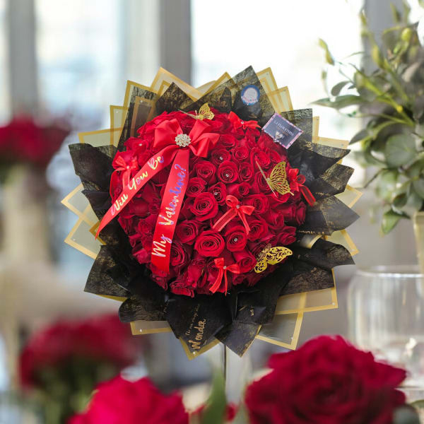 Round bouquet of tightly packed red roses wrapped in black and gold paper with red ribbons.