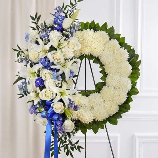 Standing white and blue floral wreath with roses, lilies, and mums on a metal easel