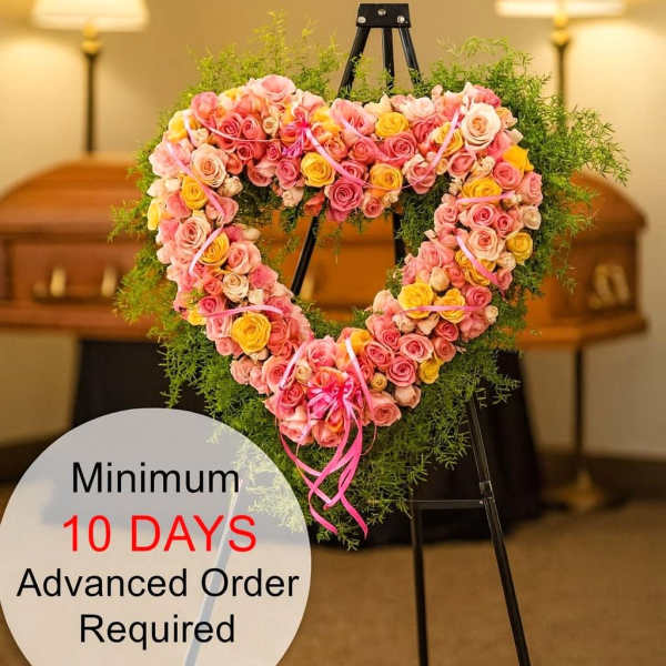 Heart-shaped wreath of pink and yellow roses on a metal easel in a funeral setting.