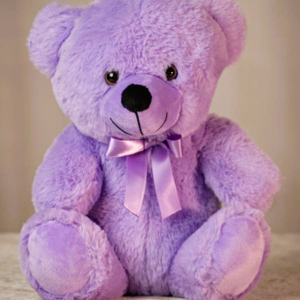 Lavender plush teddy bear with a satin bow sitting against a neutral background.