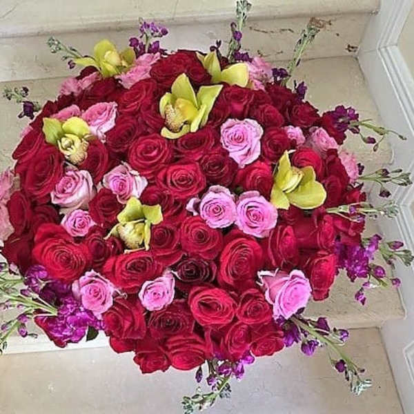 Dense round arrangement of red and pink roses accented with yellow orchids and small purple flowers