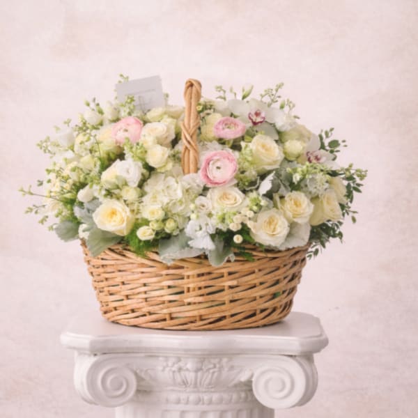Basket arrangement of cream roses with soft pink ranunculus and white blooms on a white pedestal.
