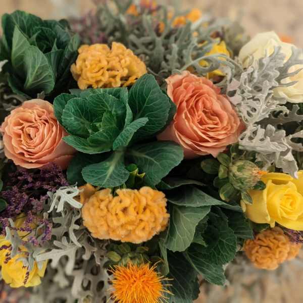 Overhead view of a mixed arrangement with peach and yellow roses, fuzzy yellow blooms, and ornamental cabbage.