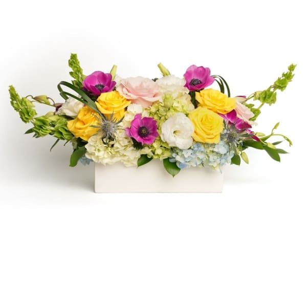 Low rectangular arrangement of yellow roses, pink anemones and mixed blooms in a white vase