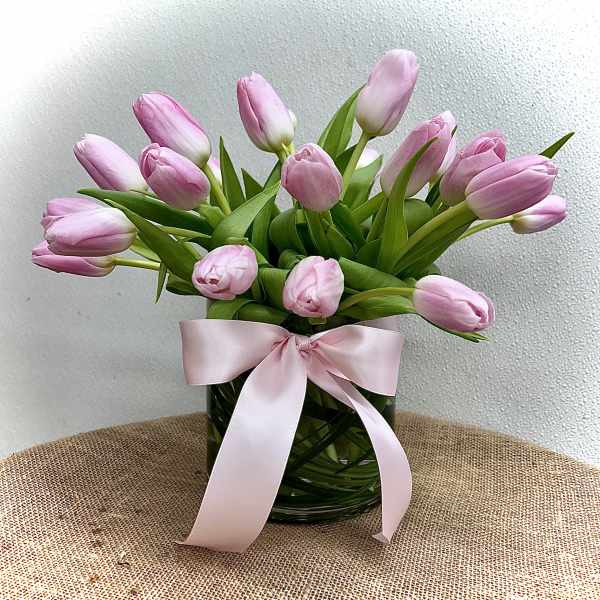 Arrangement of pink tulips in a clear glass vase wrapped with leaves and tied with a pink ribbon.