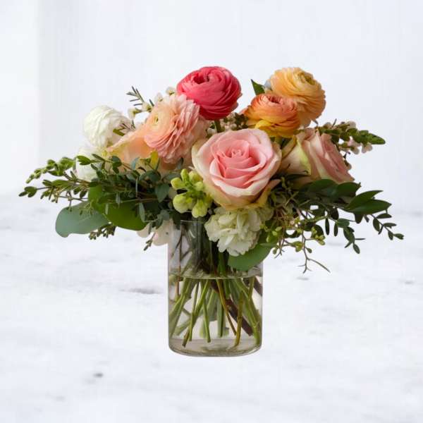 Short arrangement of pink roses and peach, white, and yellow ranunculus in a clear glass vase