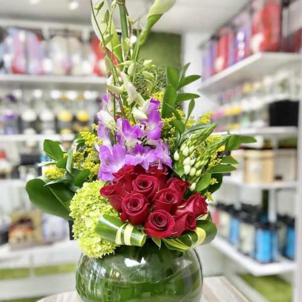 Tall arrangement with white calla lilies, purple orchids, and red roses in a round green-lined glass vase