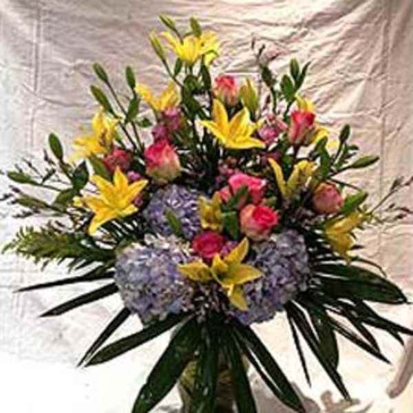 Tall arrangement of yellow lilies, pink roses, and pale lavender hydrangeas in a glass vase