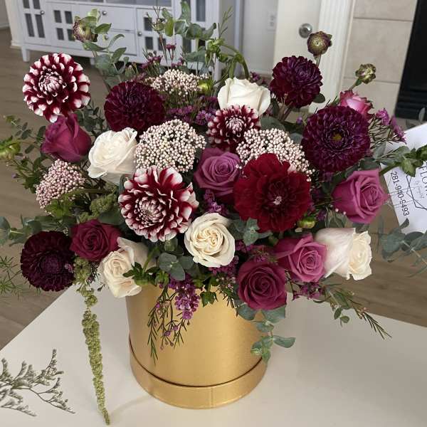 Burgundy dahlias and pink and cream roses arranged in a gold cylinder box.