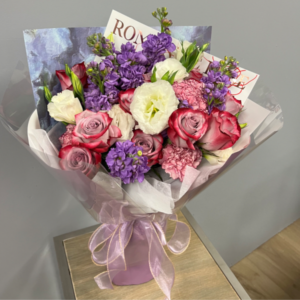 Hand-tied bouquet of pink roses, purple flowers, and white blooms wrapped in pink paper with a sheer ribbon.