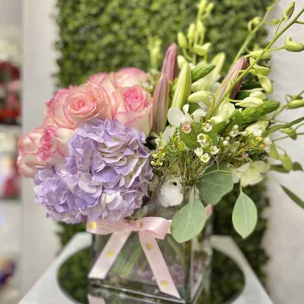 Pastel arrangement of pink roses, lavender hydrangea, and lilies in a glass cube vase with a pink ribbon.