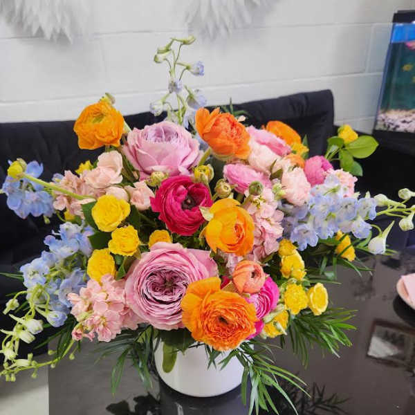 Bright mixed arrangement of pink, yellow, orange, and blue flowers in a white vase on a dark table
