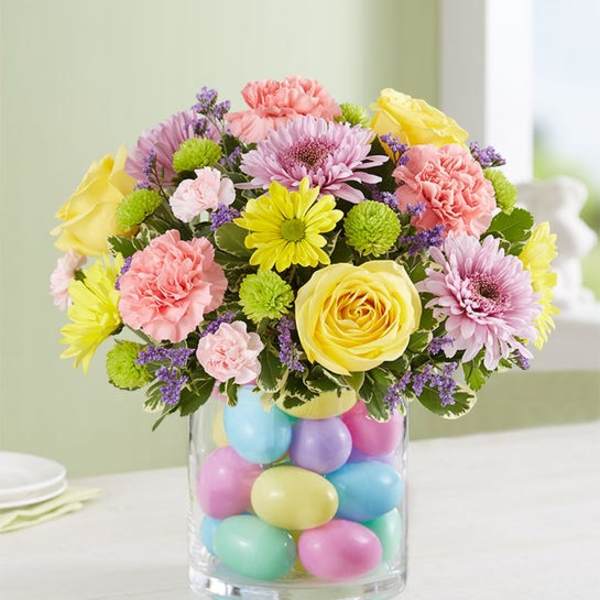 Pastel bouquet with yellow roses, pink carnations, and daisies in a glass vase filled with colorful eggs.