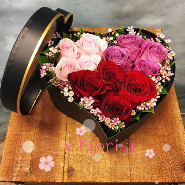 Heart-shaped black box filled with red, pink, and lavender roses and tiny pink filler flowers.