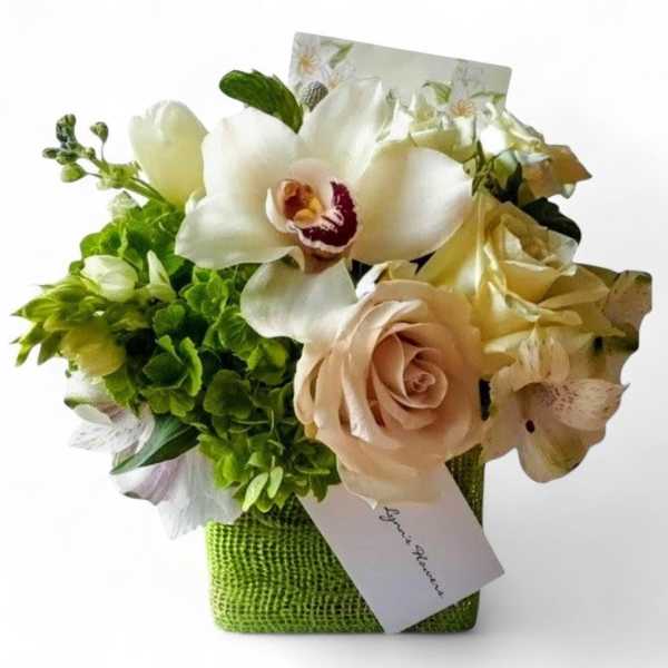 Small cube arrangement of white orchids, blush roses, and green blooms in a green vase with a card.