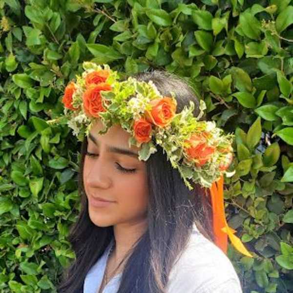 Woman wearing an orange rose flower crown with greenery and ribbon, standing in front of a leafy hedge.