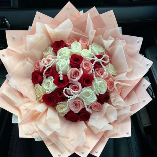 Round bouquet of red, pink, and white roses wrapped in layered peach paper with pearl accents.